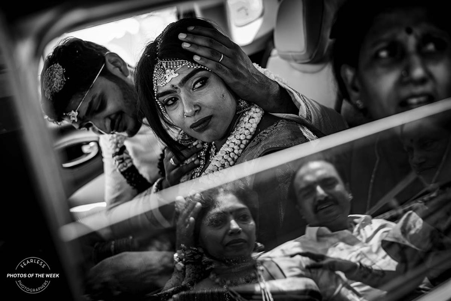 Black-and-white photograph of a groom resting his head against his bride’s inside a car, with people reflected in the window glass, selected for Fearless Photographers’ ‘Photos of the Week’