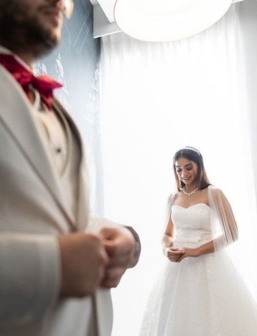 Theresa in her white wedding gown beside a bright window, Geet in a white suit in the foreground