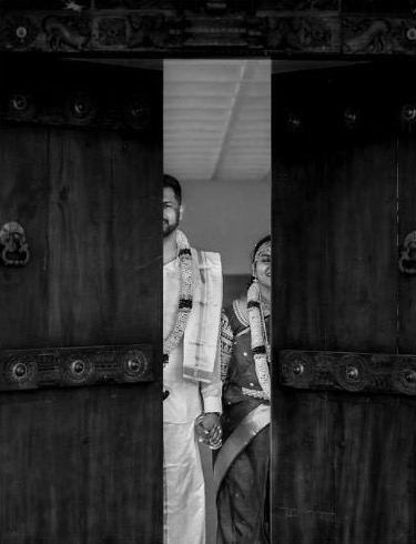 Pooja and Jagdheep peeking through a carved wooden temple door, in black and white