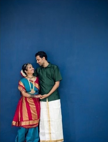 Pooja and Sajith smiling at each other against a deep blue wall, she in a red and blue silk saree, he in a green shirt and white veshti